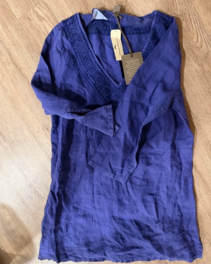 Casual phone photo of a blue embroidered tunic, wrinkled on a wood floor with the price tag still attached